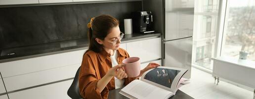 portrait de Jeune femme profiter sa fin de semaine, en train de lire une livre et en buvant thé à maison, portant des lunettes et décontractée vêtements photo