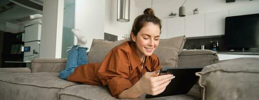 fin de semaine et mode de vie concept. Jeune femme mensonge sur canapé avec numérique tablette, défilement social médias, en train de lire ebook ou en train de regarder la télé séries sur app photo