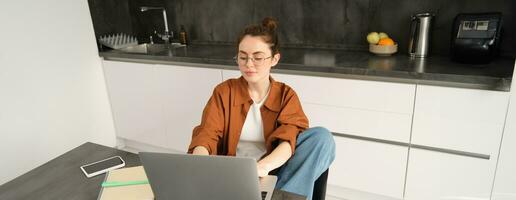 portrait de Jeune femme, étudiant dans cuisine, séance avec ordinateur portable, travail de maison, en train d'étudier ou Faire devoirs, connecte à en ligne bavarder ou cours de ordinateur photo