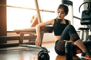 jeune femme asiatique relaxante dans une salle de fitness et un centre de club de sport avec équipement et haltère. concept d'entraînement et de musculation. thème de la beauté et de la santé. fond de gymnase photo