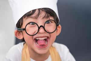 portrait mignon petit garçon heureux asiatique faisant le visage de la langue drôle et intéressé par la cuisine dans la cuisine à domicile. les modes de vie et la famille des gens. concept d'ingrédients alimentaires faits maison. cuisson du gâteau de noël et cuisinier photo