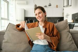 souriant Jeune brunette femme, est assis sur canapé dans vivant chambre, détient carnet de notes, lit sa Remarques, études pour examen, étudiant Est-ce que sa devoirs à Accueil photo