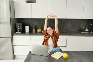 productivité et mode de vie concept. magnifique Jeune femme finitions fonctionnement, élongation mains avec satisfait expression, séance dans cuisine avec portable et documents, a éloigné lieu de travail à Accueil photo