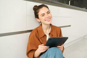 Jeune entrepreneurs. souriant mignonne femme avec numérique tablette, séance avec gadget sur cuisine sol et à la recherche content, en train d'étudier à distance, en train de lire sur sa dispositif photo