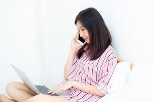 jeune femme asiatique assise sur le lit à l'aide d'un ordinateur portable et parlant avec un téléphone intelligent. photo