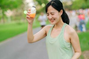 femelle joggeur. en forme Jeune asiatique femme avec vert tenue de sport en buvant biologique Orange jus après fonctionnement et profiter une en bonne santé Extérieur. aptitude coureur fille dans Publique parc. bien-être étant concept photo