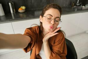 selfie de content Jeune et insouciant femme, prise photo sur mobile téléphone avec élargi main, posant et souriant, séance dans cuisine dans des lunettes