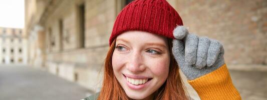 photo du visage de content roux fille avec taches de rousseur, porte rouge chapeau et gants dans hiver, des promenades autour ville sur froid temps et sourit