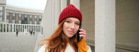 mobile haut débit et personnes. souriant Jeune roux femme des promenades dans ville et pourparlers sur mobile téléphone, appel ami sur téléphone intelligent, en utilisant l'Internet à faire une appel à l'étranger photo