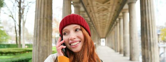 souriant mignonne roux femme fait du une téléphone appel, détient Téléphone près année, a mobile conversation, en utilisant téléphone intelligent sur rue photo