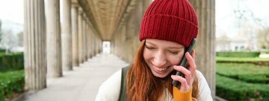 portrait de roux européen fille dans rouge chapeau, fait du une téléphone appel, des promenades dans ville et pourparlers à ami sur téléphone intelligent photo