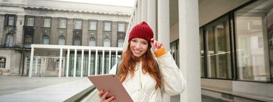 élégant gingembre fille, touristique des promenades avec numérique tablette autour ville, femme connecte à internet sur sa gadget, à la recherche en haut information, envoyer des SMS message photo