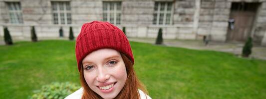 verticale portrait de mignonne roux femelle étudiant dans rouge chapeau et chaud gants, est assis dans parc sur banc, sourit et regards mignonne à caméra photo