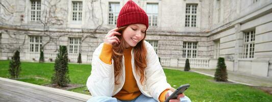 Jeune souriant roux fille est assis sur banc et les usages téléphone intelligent application, lit nouvelles en ligne, montres vidéo sur mobile téléphone tandis que détendu dans parc photo