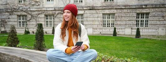 Jeune souriant roux fille est assis sur banc et les usages téléphone intelligent application, lit nouvelles en ligne, montres vidéo sur mobile téléphone tandis que détendu dans parc photo