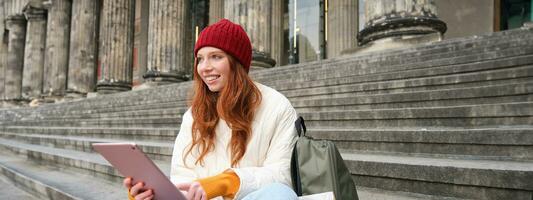 magnifique Jeune moderne fille avec rouge cheveux, détient numérique tablette, est assis sur escaliers près musée et connecte Publique l'Internet, envoie message sur gadget app photo