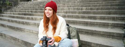 Jeune étudiant, photographe est assis sur rue escaliers et chèques sa coups sur professionnel caméra, prise Photos en plein air