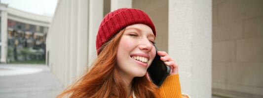 mobile haut débit et personnes. souriant Jeune roux femme des promenades dans ville et pourparlers sur mobile téléphone, appel ami sur téléphone intelligent, en utilisant l'Internet à faire une appel à l'étranger photo