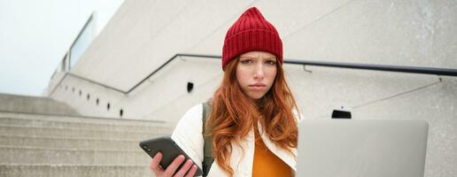 portrait de roux femme est assis sur escaliers, les usages portable et détient téléphone intelligent, regards confus et dérangé à ordinateur filtrer, essaie à relier à Wifi Publique l'Internet photo