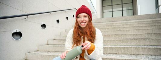 souriant voyageur, roux fille touristique est assis sur escaliers avec ballon, les boissons chaud café de thermos tandis que en voyageant et tourisme autour étranger ville, est assis sur escaliers et repose photo