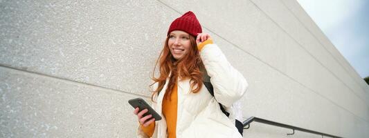 élégant roux femme dans rouge chapeau, penche sur mur sur rue, détient téléphone intelligent, les usages l'Internet téléphone application, lit message photo