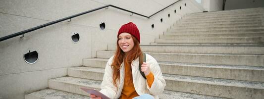 content roux femme dans rouge chapeau, est assis sur escaliers en plein air, les usages tablette, connecte à Wifi près Publique bâtiment, lit numérique livre tandis que attend sur rue photo