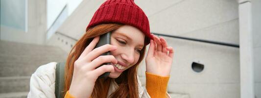 Jeune élégant roux fille dans rouge chapeau, est assis sur rue et pourparlers sur mobile téléphone, a Téléphone conversation, anneaux sa ami tandis que se détend en plein air photo