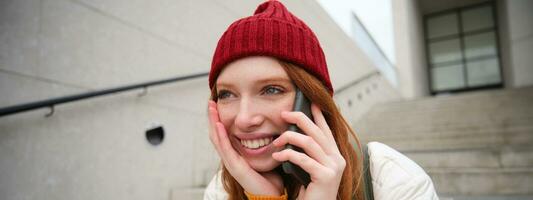 Jeune élégant roux fille dans rouge chapeau, est assis sur rue et pourparlers sur mobile téléphone, a Téléphone conversation, anneaux sa ami tandis que se détend en plein air photo