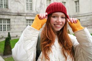 Jeune souriant roux femme en marchant dans magnifique ville attractions, portant sac à dos, rouge chapeau et manteau, à la recherche autour avec content visage photo