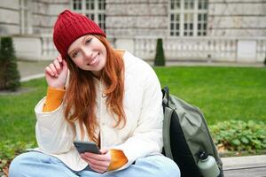 Jeune souriant roux fille est assis dans parc avec téléphone intelligent, envoyer des SMS message sur mobile téléphone, en utilisant Téléphone, de liaison à Publique Wifi et surfant l'Internet photo