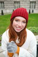 verticale portrait de mignonne roux femelle étudiant dans rouge chapeau et chaud gants, est assis dans parc sur banc, sourit et regards mignonne à caméra photo