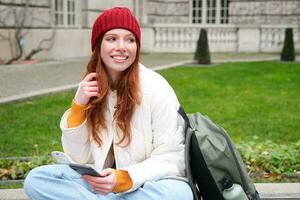 roux fille, femelle étudiant est assis avec mobile téléphone sur banc dans parj, penche sur sa sac à dos. femme navigation social médias app alimentation sur sa téléphone intelligent photo