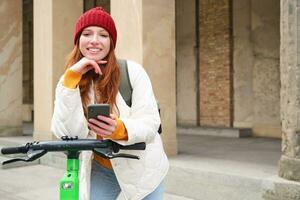 souriant Jeune roux femme avec mobile téléphone, les loyers escooter en utilisant téléphone intelligent application, équitation autour ville, Urbain transport et gens concept photo