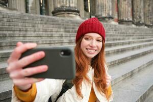 Jeune roux touristique prend selfie dans de face de musée sur escaliers, détient téléphone intelligent et regards à mobile caméra, fait du photo de se avec téléphone