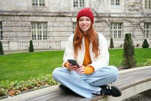 portrait de élégant Jeune femme, 25 années, est assis sur banc dans parc et les usages mobile téléphone, lit en ligne nouvelles, messages ou montres vidéo sur téléphone intelligent application, connecte à Publique Wifi photo