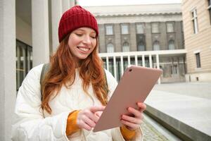 content roux fille dans rouge chapeau, des promenades autour ville avec numérique tablette, connecte à Publique l'Internet Wifi et regards pour itinéraire, regards à carte sur sa gadget photo