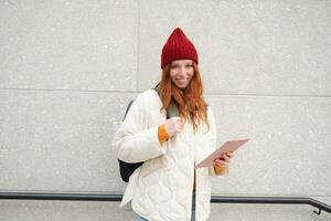 Jeune roux femme avec rouge chapeau, les usages sa numérique tablette en plein air, des stands sur rue avec gadget, connecte à Wifi l'Internet et recherches pour une emplacement dans l'Internet photo