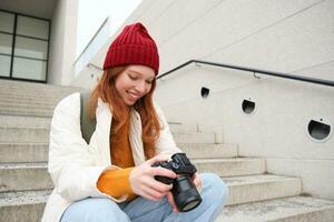 souriant roux fille photographe, chèques sa coups, détient caméra et regards à filtrer, prend Photos en plein air, des promenades autour rue et Est-ce que style de rue tournage