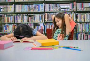 bonheur deux jolies filles de la diversité lisant un livre et taquinant pour frapper un ami endormi dans la bibliothèque de l'école drôle. mode de vie, éducation et amitié des gens. enfants et enfants activité de groupe loisirs photo