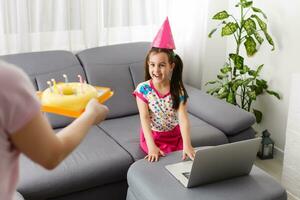 les enfants virtuel anniversaire fête avec gâteau en ligne ensemble avec sa ami dans vidéo conférence. avec numérique Téléphone pour une en ligne réunion. fille célébrer anniversaire en ligne dans quarantaine temps photo