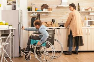 femme dans cuisine à la recherche à mari avec en marchant invalidité en essayant à ouvert réfrigérateur porte. désactivée paralysé handicapé homme avec en marchant invalidité en intégrant après un accident. photo