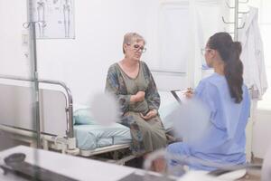 retraité femme pendant médical vérifier avec infirmière dans hôpital bureau. assistant dans bleu uniforme prise Remarques sur presse-papiers. photo