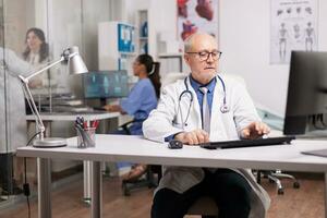 personnes âgées vieilli médecin en utilisant ordinateur dans hôpital cabinet tandis que Jeune médecin parlant avec femelle patient sur clinique couloir et infirmière dactylographie rapport sur pc. photo