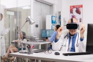 Sénior médecin portant virtuel réalité casque dans hôpital Bureau tandis que Jeune thérapeute est discuter avec désactivée femme dans fauteuil roulant et infirmière dans bleu uniforme. photo