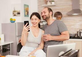 de bonne humeur couple parlant avec copains sur vidéo appel dans le Matin séance sur le chaise dans le cuisine. joyeux marié mari et épouse fabrication marrant visages tandis que prise une photo pendant petit déjeuner dans cuisine.