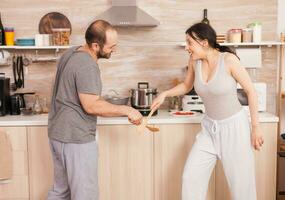 content Jeune couple en jouant avec cuillères dans le cuisine. insouciant épouse et mari en riant ayant amusement marrant profiter la vie authentique marié gens positif content relation photo