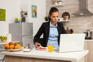 affaires femme en mangeant petit déjeuner et travail sur portable. concentré affaires femme dans le Matin multitâche dans le cuisine avant Aller à le bureau, stressant façon de vie, carrière et buts à rencontrer photo