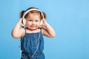 mignonne peu fille dans écouteurs écoute à la musique en utilisant une tablette et souriant sur bleu Contexte photo