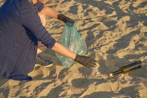 Jeune bénévole dans noir gants est en marchant avec des ordures sac le long de une sale plage de le rivière et nettoyage en haut poubelle. gens et écologie. fermer. photo