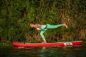 Jeune femme sont Faire yoga sur une supporter en haut pagayer planche souper sur une magnifique Lac ou rivière photo